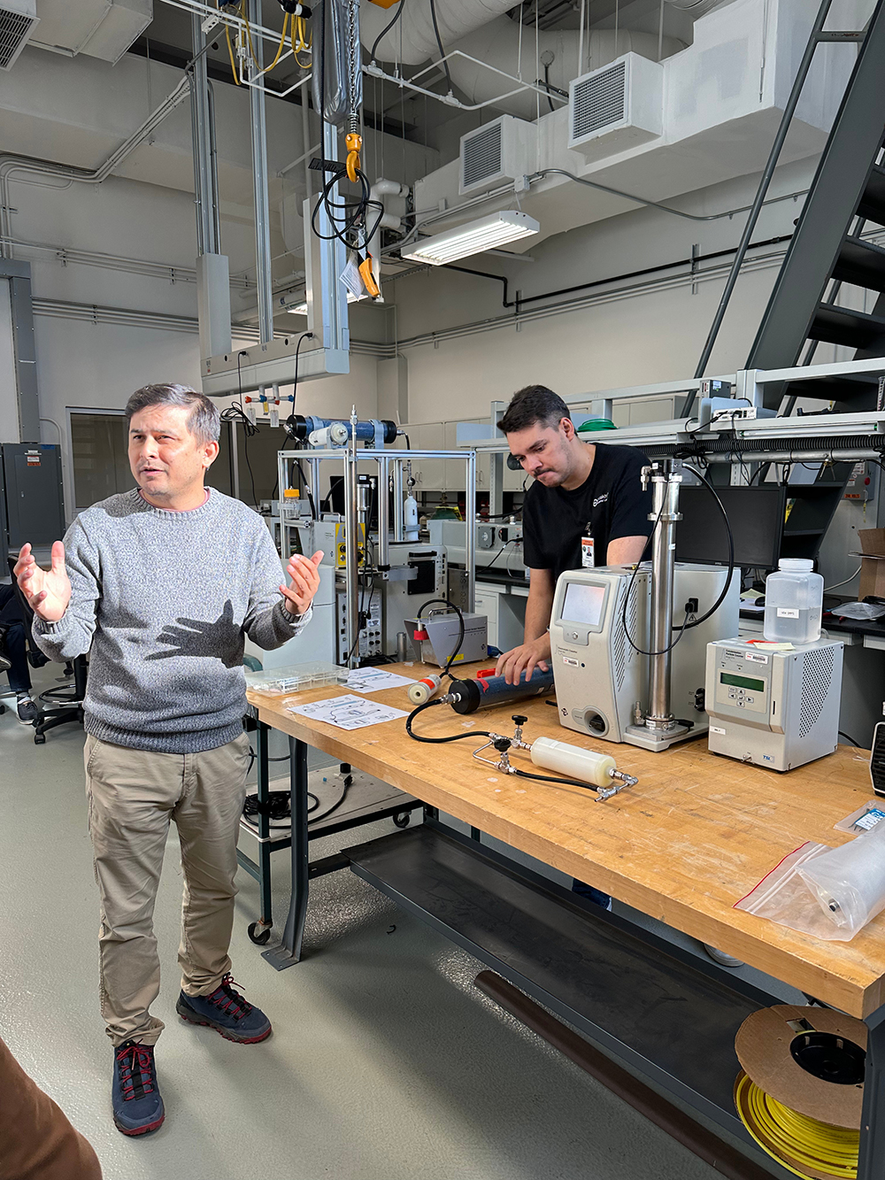 Ashish Singh gestures with his hands while Delano Campos de Oliveira stands behind a table and works with a scanning mobility particle sizer calibration system. Photo is by Olga Mayol-Bracero, Brookhaven National Laboratory.