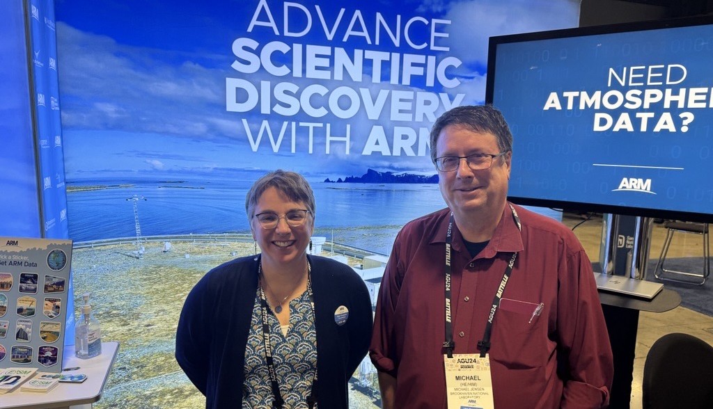Mike Jensen and Jen Delamere stand in front of a large exhibit and giant screen. Next to Jen is a table with a placard and ARM and DOE stickers.